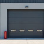Modern dark grey sectional garage door with small windows. Industrial building facade features corrugated metal siding. Red bollards mark entrance.