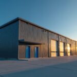Modern warehouse building exterior with metal exterior at sunset. Industrial commercial property with large garage roller doors, blue entrance. Storage facility small distribution center stands against