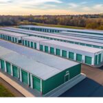 Aerial view of green and white self-storage buildings surrounded by trees under clear sky.