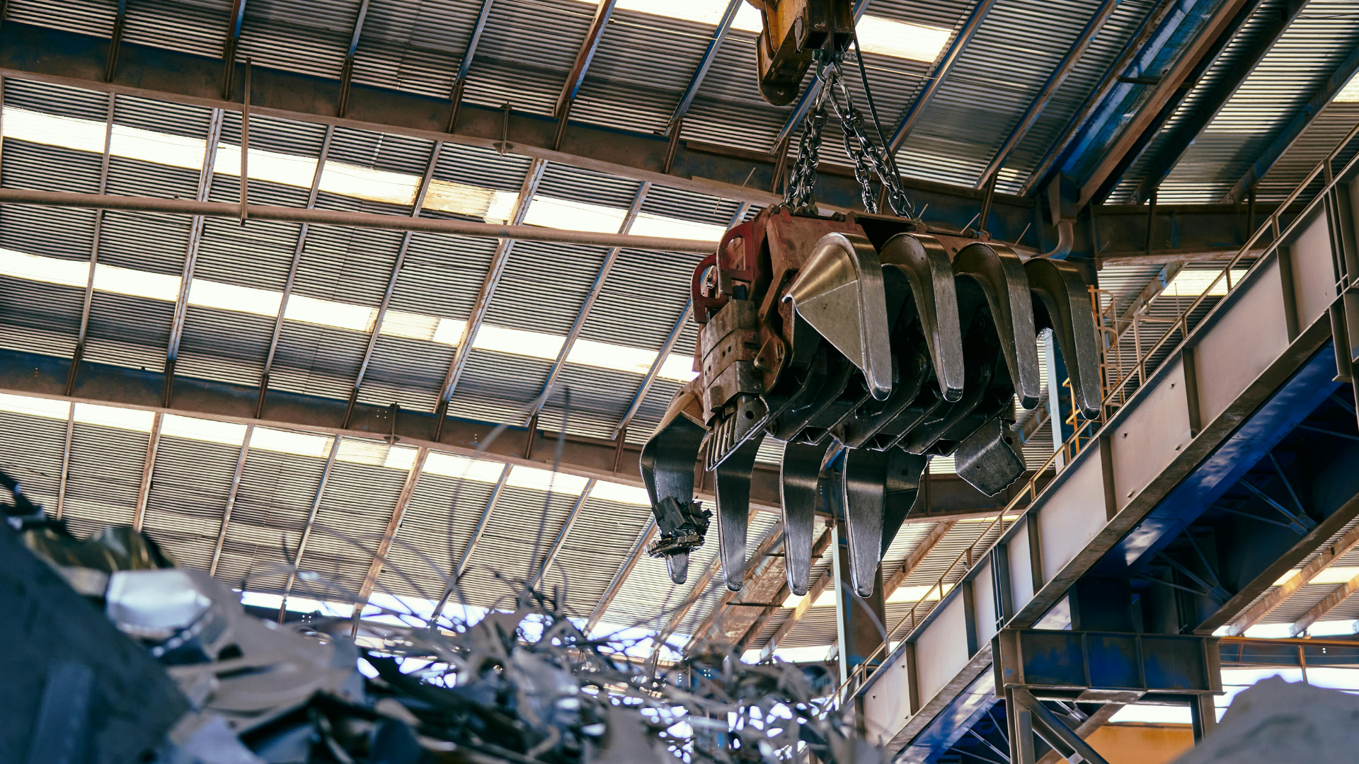 Industrial scrap processing facility with an overhead crane and metal grapple handling recycled steel inside a steel-framed warehouse.