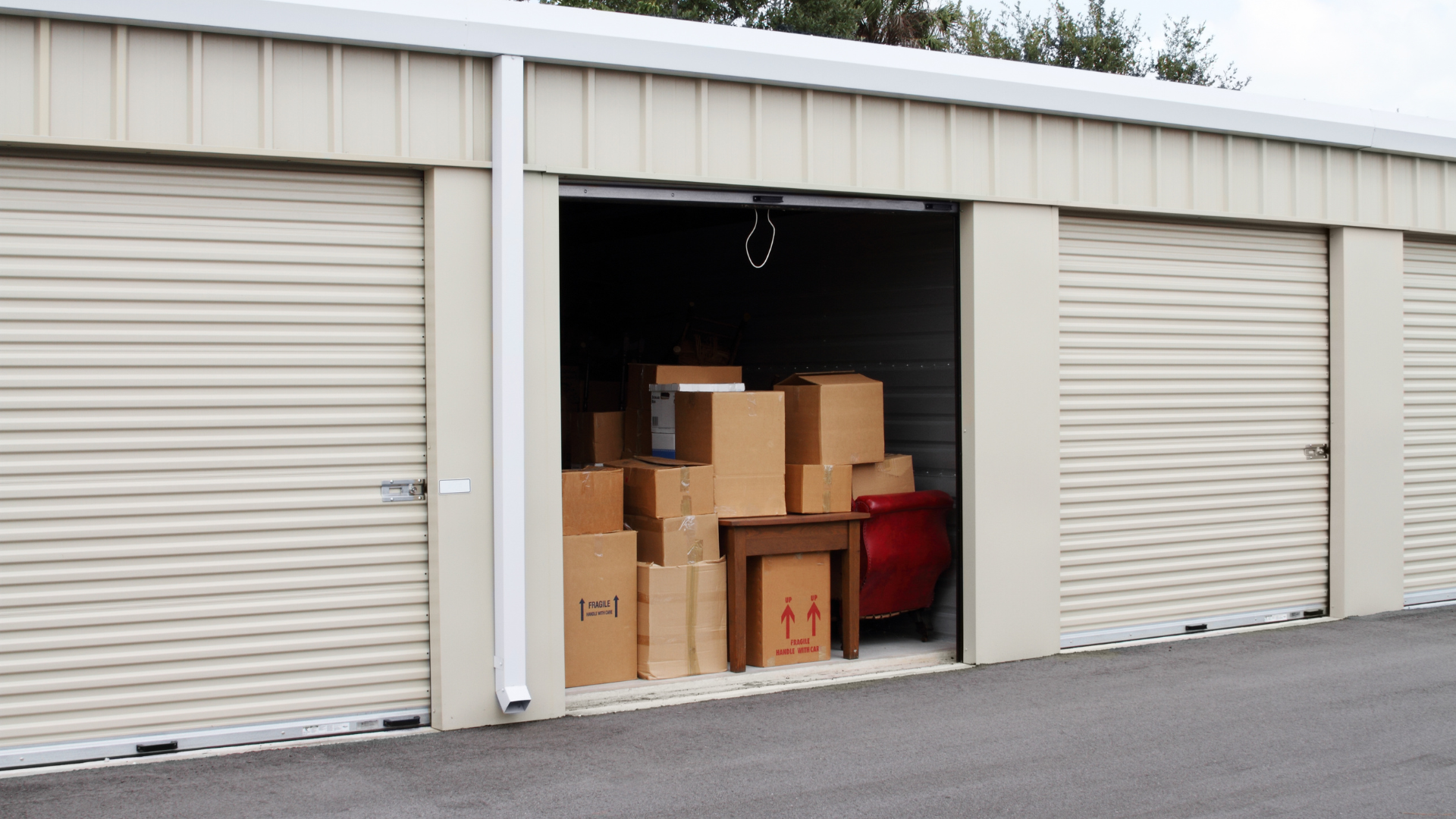 Self-storage facility with roll-up unit doors, including an open storage unit filled with moving boxes and household furniture.
