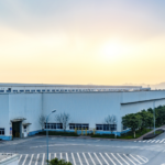 Large industrial manufacturing and warehouse complex with wide access roads, landscaped surroundings, and expansive production buildings at sunrise.